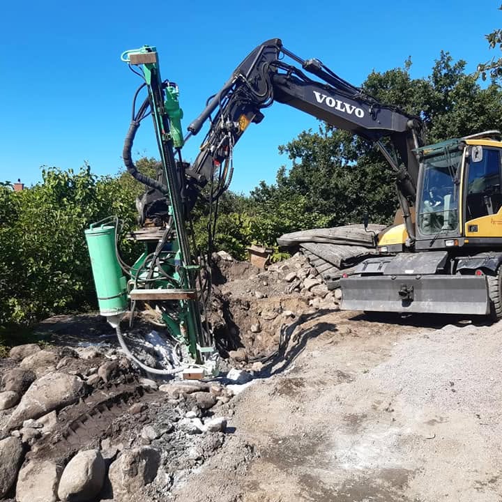 Volvo-grävmaskin med borrigg som bergborrar tätt intill en husgrund